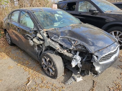 Damaged black Kia sedan with extensive front-end wreckage parked among other cars. The vehicle's condition highlights a recent accident.