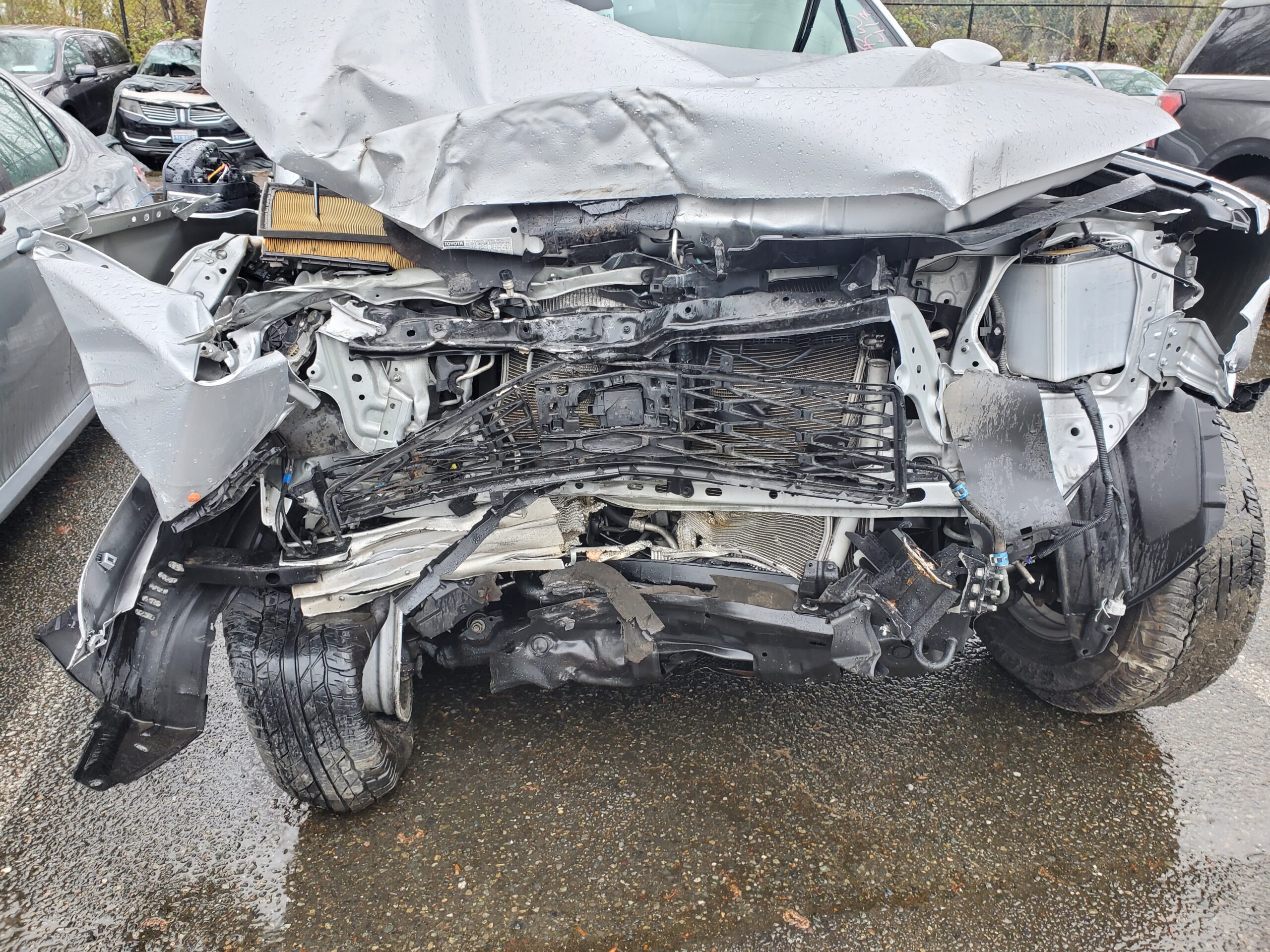 Severely damaged front end of a silver vehicle, exposing wires and components, indicating a significant collision. Rain-soaked pavement reflects the wreckage.