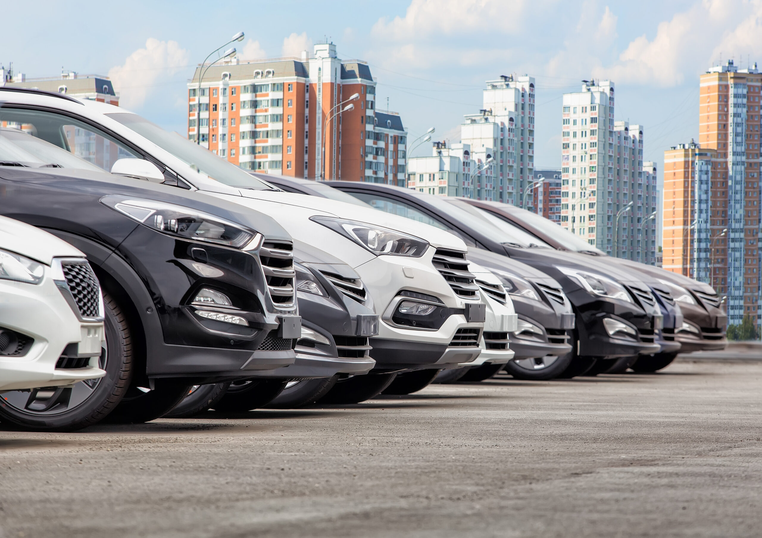 A row of parked cars in varying colors, with modern high-rise buildings in the background under a partly cloudy sky.